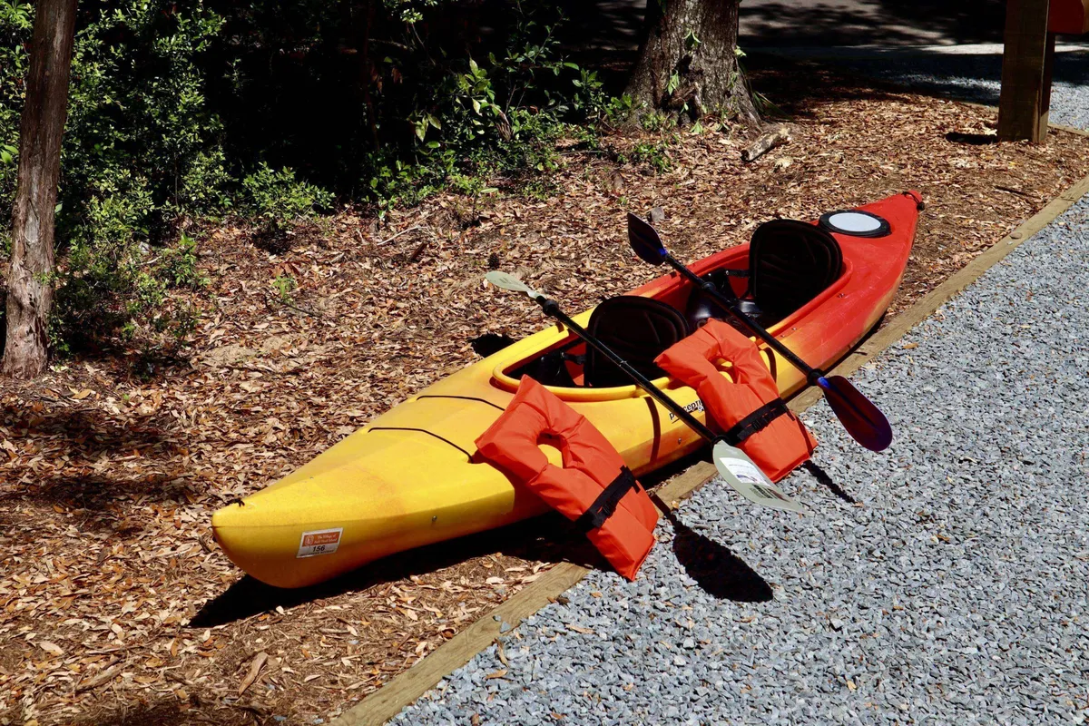 This vibrant exterior image features a yellow kayak and a red kayak laid out with paddles and life jackets, inviting immediate use for water adventures. The setting highlights excitement and readiness for outdoor fun.