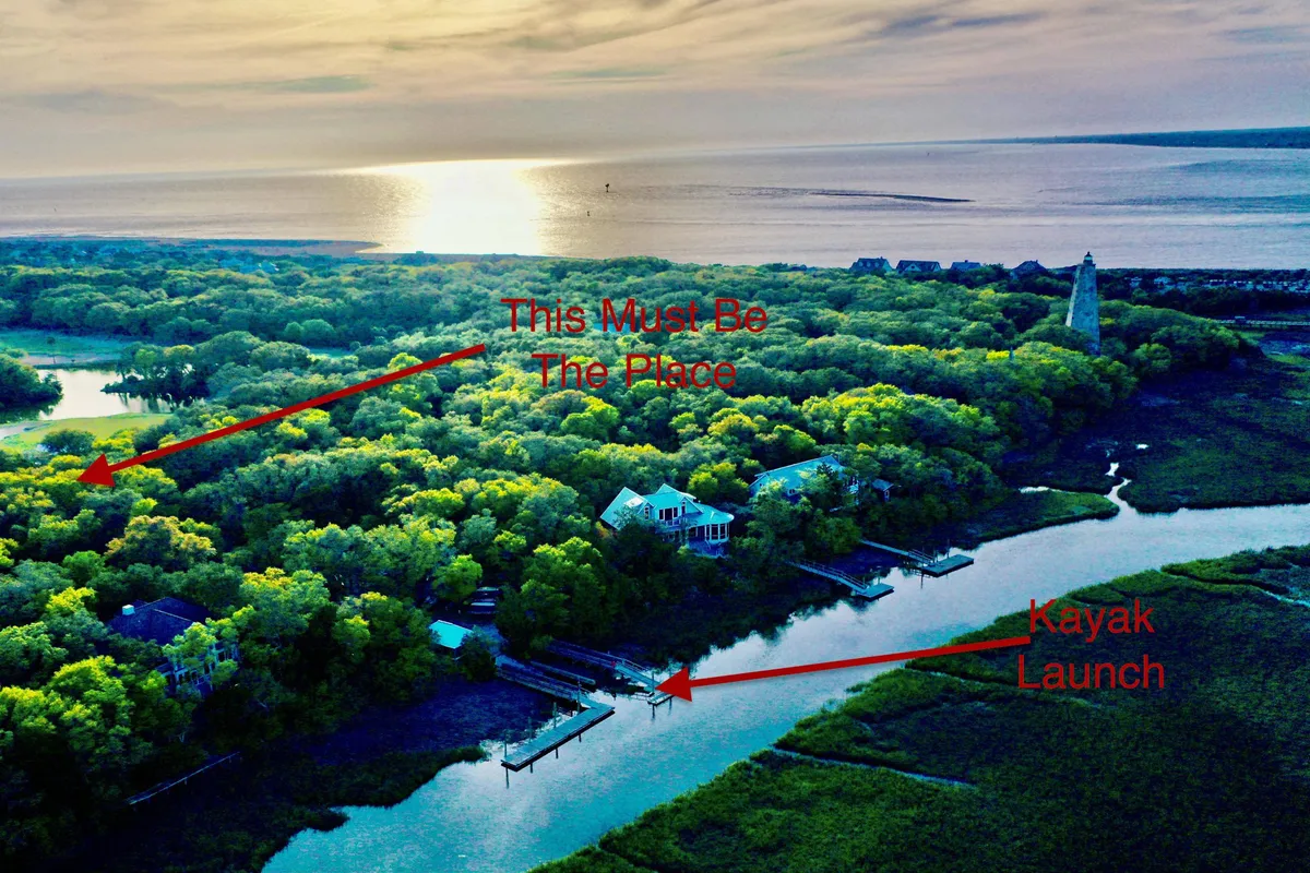 This exterior image showcases lush green trees flanking houses by the water, a kayak launch in the foreground, a distant lighthouse, and a beautiful sunset over the water. The scene captures kayaks at the ready and the immediate access to aquatic adventure.