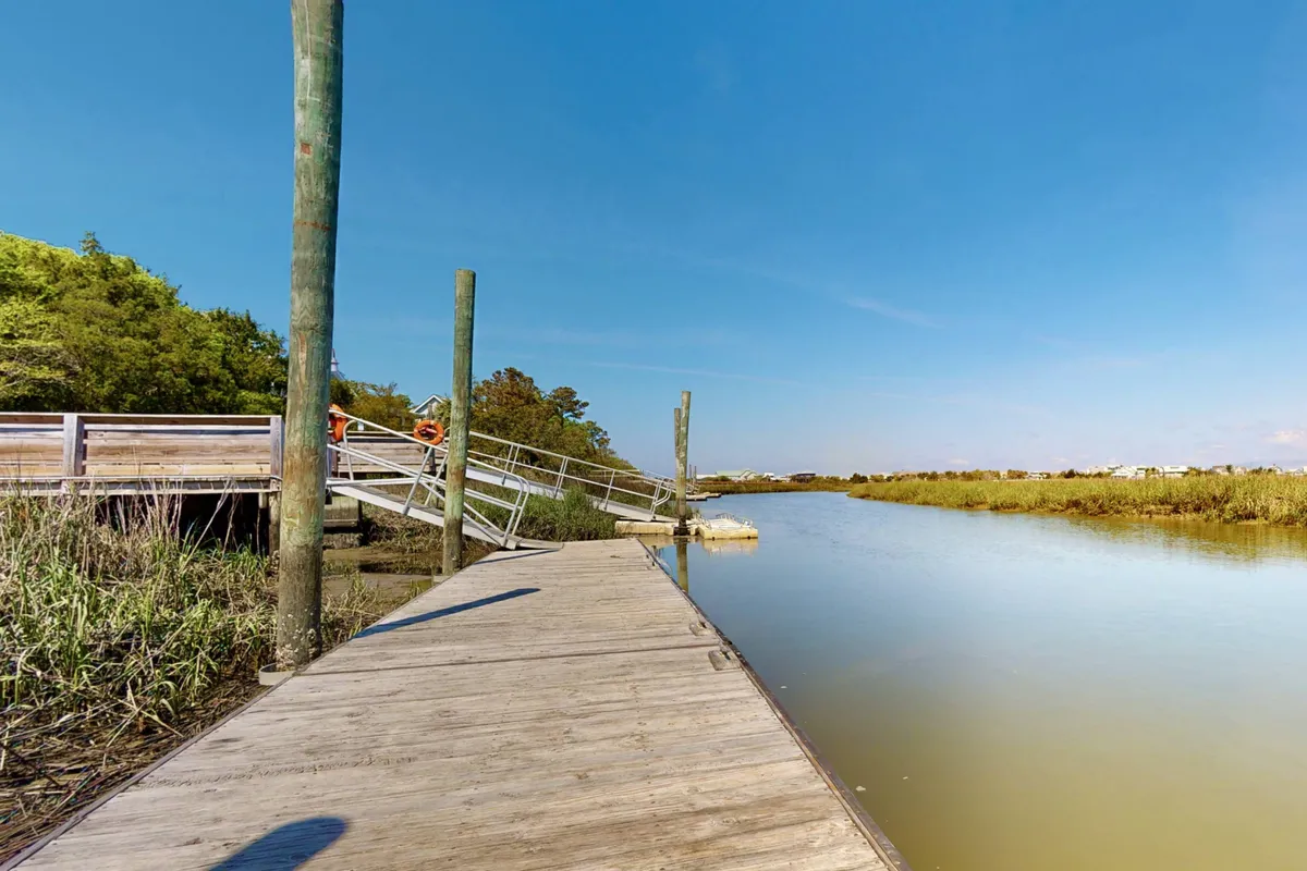 A peaceful exterior shot depicting a wooden dock stretching over calm water, surrounded by lush greenery and beneath a clear blue sky. The scene suggests an inviting spot for relaxing or launching into water-based adventures.