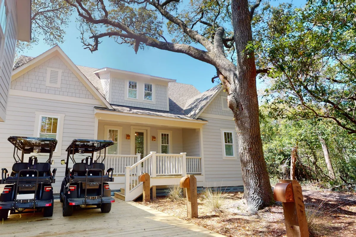 The image centers on the house entrance, with two golf carts parked under a large tree. The porch hints at a welcoming interior while the golf carts are prominently displayed, ready for island exploration.