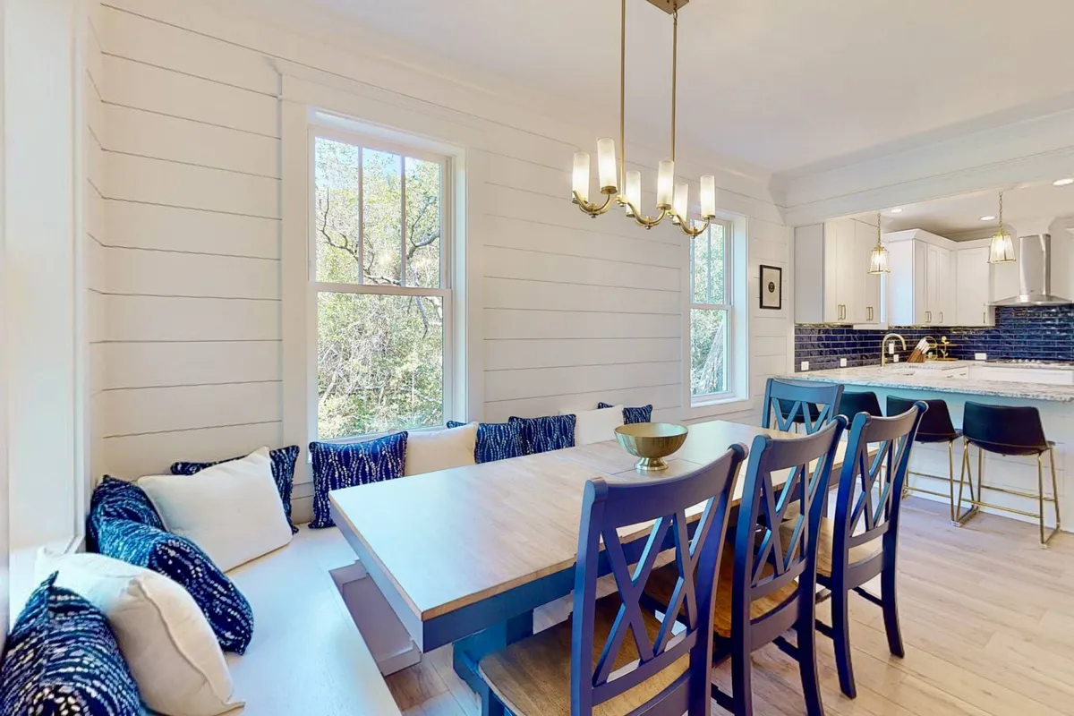 A warm dining area with a central table, window seating, elegant chandeliers, and a view into the adjacent kitchen. The cozy layout is ideal for family gatherings, sharing stories, and enjoying meals together, with natural light pouring in to enhance the welcoming mood.