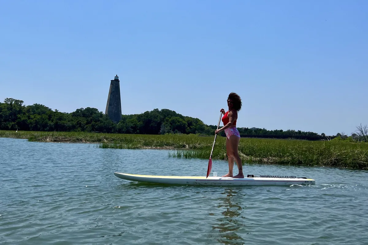 This image features a paddleboarder gliding across calm waters with the historical lighthouse and lush greenery framing the background. Water reflections and the open marshland create a sense of adventure and serenity.