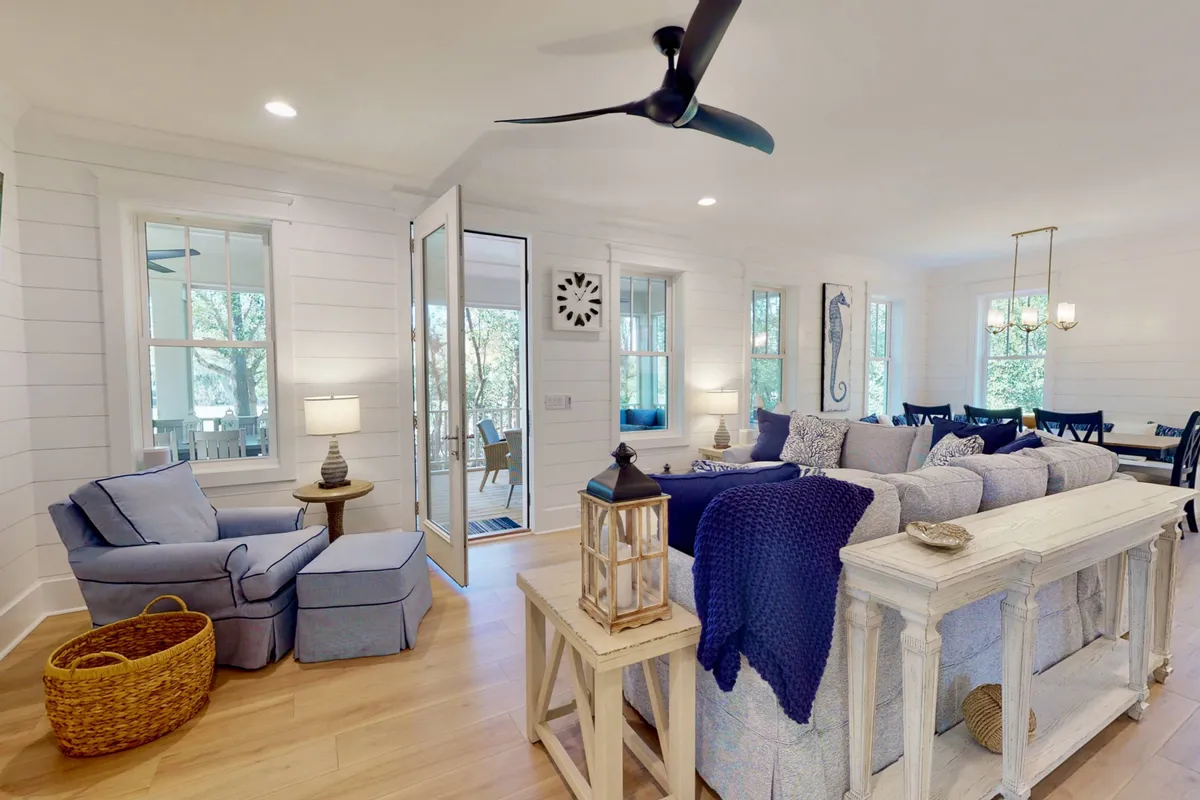 This image showcases an open and airy living space featuring a large sectional sofa, a cozy blue armchair, and a dining area. The open door leads directly to an outdoor space, emphasizing the seamless indoor-outdoor flow. Natural light fills the room, and the arrangement of furniture invites gatherings and relaxation.