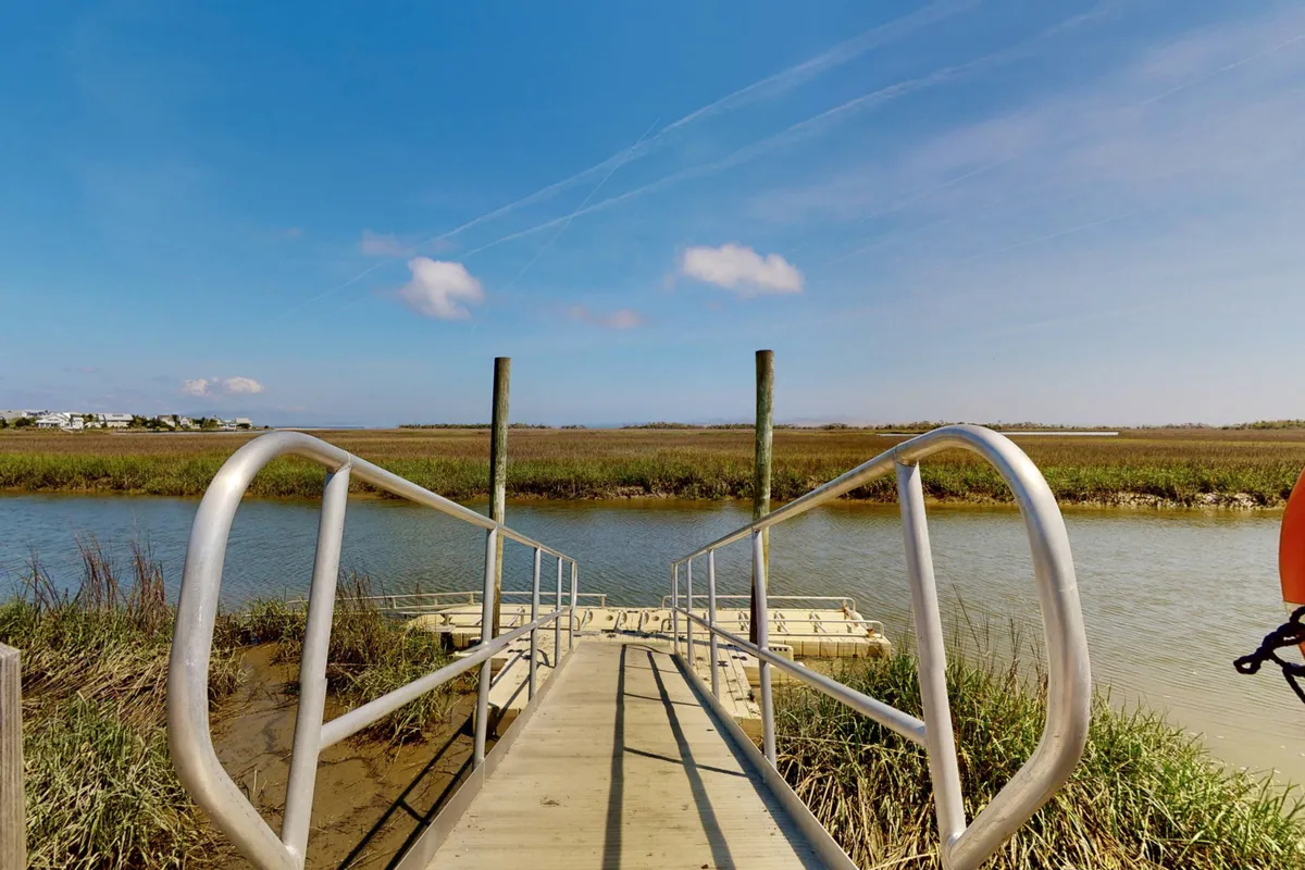 A wooden dock extends over calm marshland waters under an open sky, creating an idyllic spot for nature walks, birdwatching, and peaceful moments by the water. The surrounding marshland and natural views emphasize the island's connection to water and wildlife.