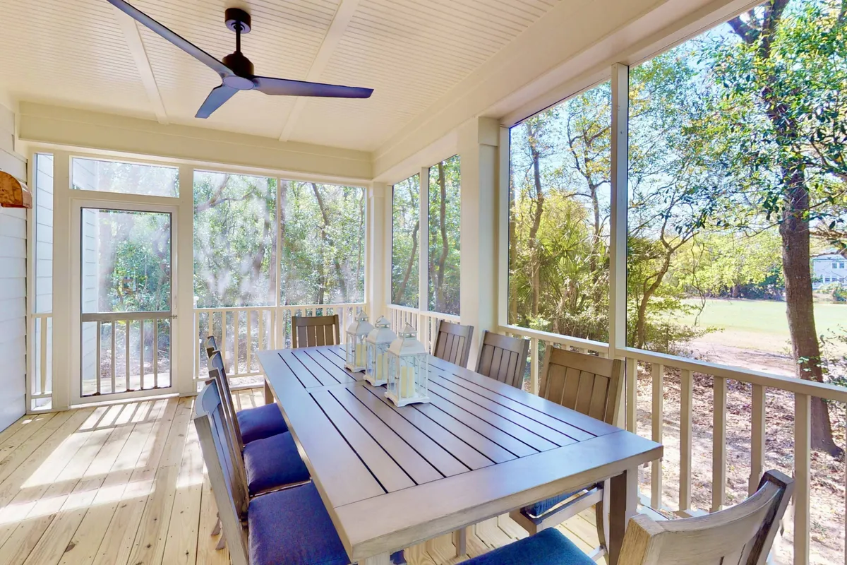 A spacious screened-in porch features a large dining table and ceiling fan, with views out through screened windows to the surrounding greenery. The setting feels tranquil and connected to nature, perfect for unwinding after time at the beach.