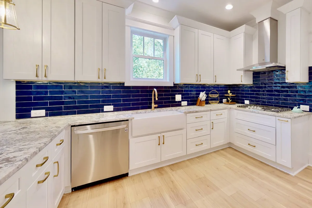 A vibrant kitchen scene featuring blue backsplash tiles, gold hardware, a granite countertop, and a sunlit kitchen window. The workspace is fresh and lively, with visible appliances and thoughtful design details that make the space both functional and joyful for group cooking and entertaining.
