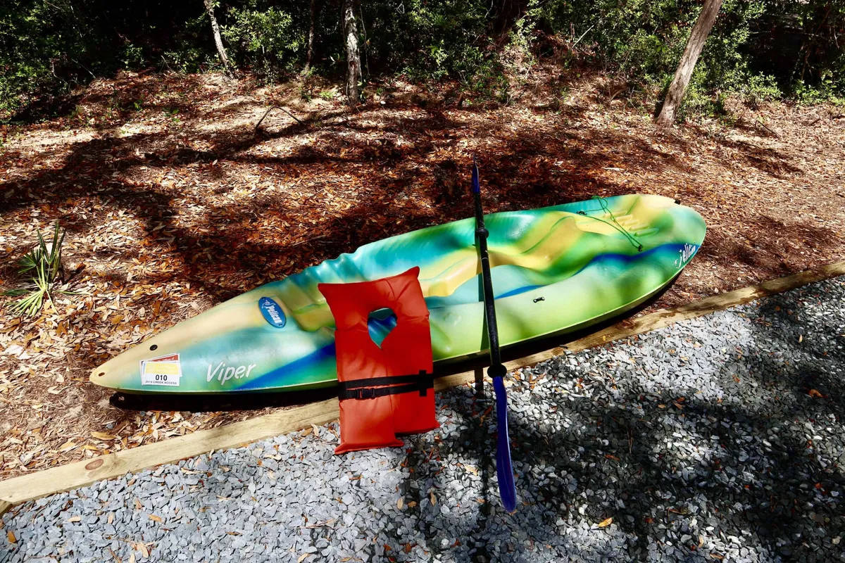 A colorful kayak, orange life vest, and paddle are ready by the water's edge, inviting guests to embark on a kayaking journey. The setting suggests both anticipation and ease of access, surrounded by peaceful natural scenery.