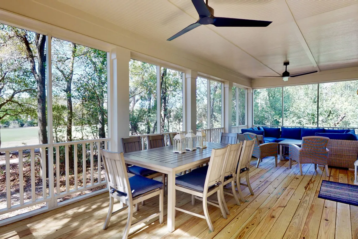 The image showcases a bright, inviting dining area with a large table perfect for gatherings, expansive windows that fill the space with natural light, and a seamless transition to outdoor seating. The setting feels airy and welcoming, ideal for family meals, socializing, or unwinding with a view of Bald Head Island.